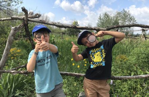 Deux garçons portant des casquettes et tenant des loupes devant leurs yeux dans un champ fleuri avec une structure en bois à l'arrière-plan