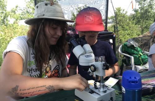 Monitrice avec tatouage au bras montrant à un jeune garçon portant une casquette rouge comment utiliser un microscope lors d'une activité éducative en plein air